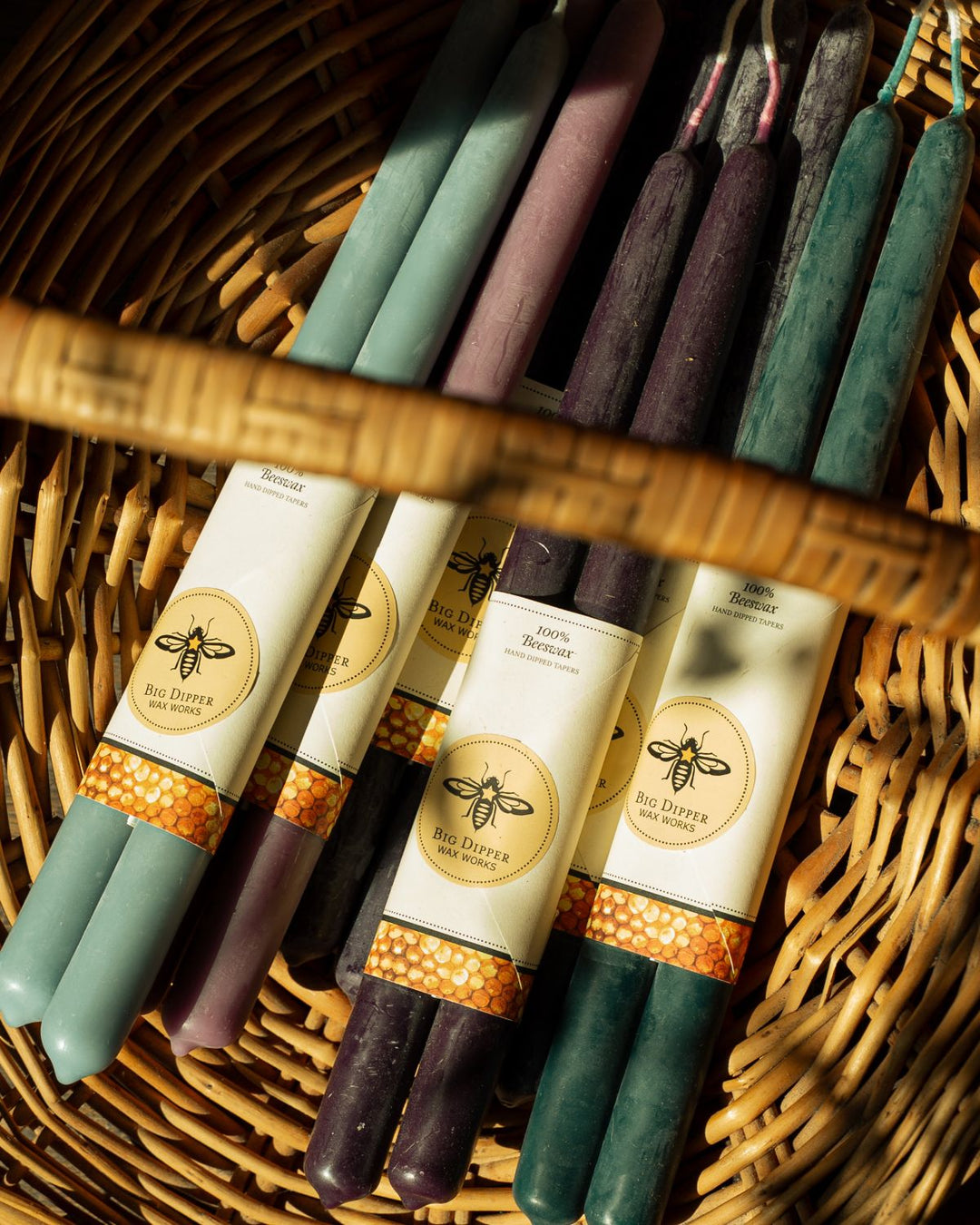 Several beeswax candles made by Synergy Herbal Works Herbal Apothecary in Tennessee in different colors, including sage green, lavender, and deep purple, wrapped in white paper labels and arranged inside a woven wicker basket.