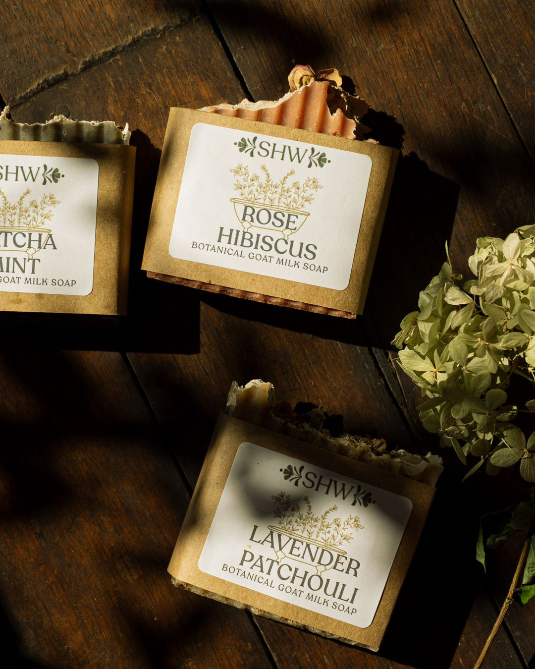 Three bars of Synergy Herbal Works botanical goat milk soap in brown kraft paper packaging, labeled Rose Hibiscus and Lavender Patchouli, arranged on a dark wooden surface with natural sunlight and shadows. All made in house by Synergy Herbal Works Herbal Apothecary in Tennessee. 