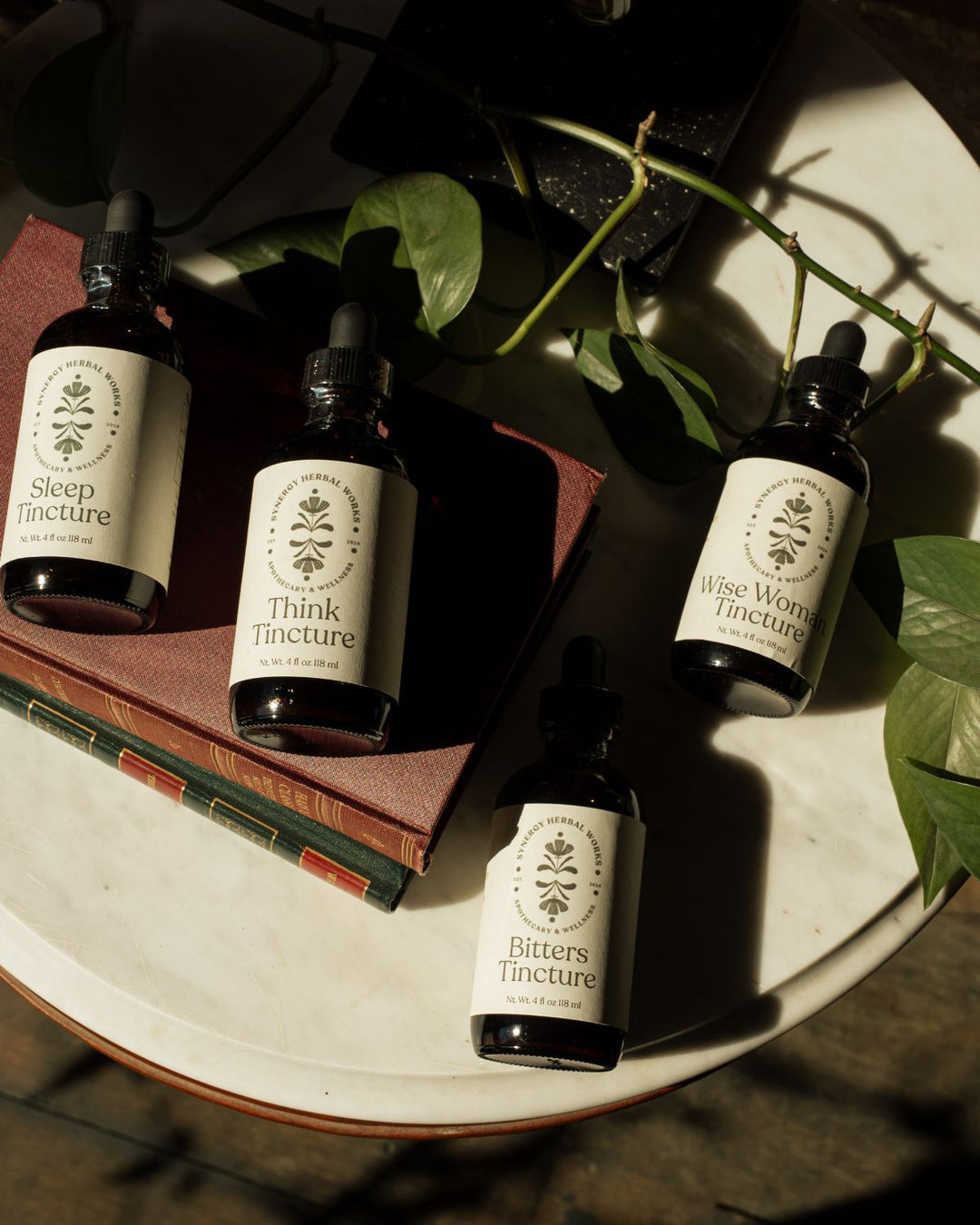 Four dark glass tinctures made by Synergy Herbal Works Herbal Apothecary in Tennessee with white labels are arranged on a white tray with books and green leaves under dramatic sunlight and shadows.