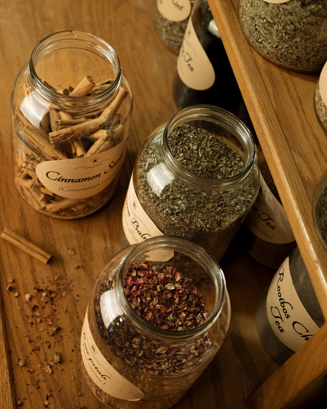 Several clear glass jars filled with loose-leaf teas and spices, including cinnamon sticks, dried herbs, and red pepper flakes, arranged on a wooden shelf and tabletop with handwritten labels all made in house by Synergy Herbal Works Herbal Apothecary in Tennessee.