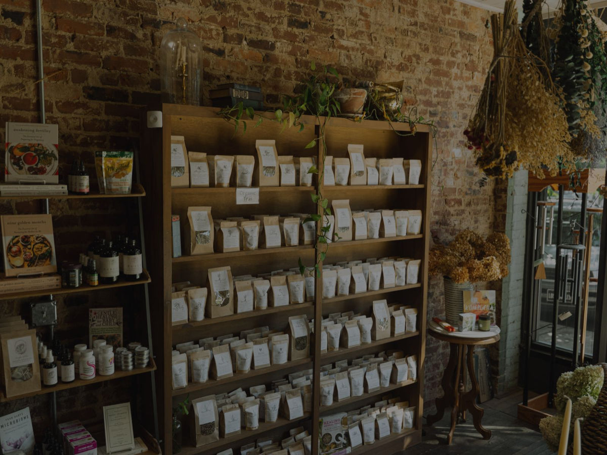 A rustic indoor herbal apothecary shop scene with a tall wooden shelving unit filled with many small white product bags, alongside other shelves of jars and packaged goods against a brick wall. Synergy Herbal Works Herbal Apothecary- The largest herbal apothecary in Tennessee. 